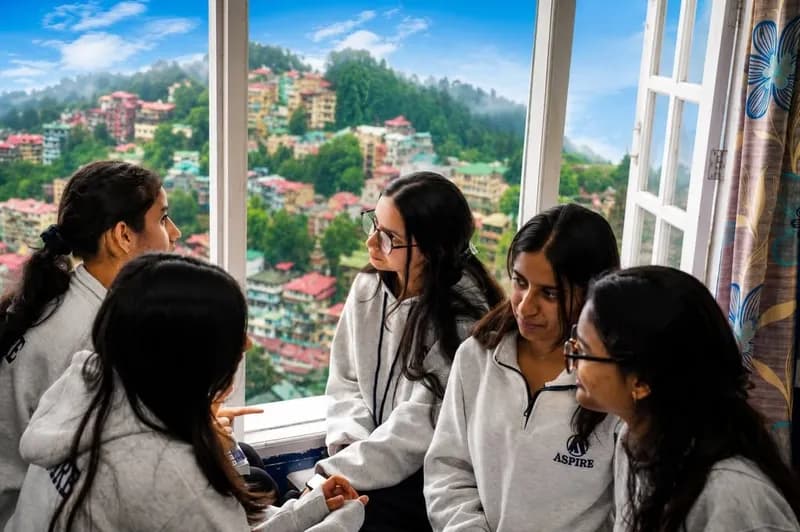 Five students in grey Aspire hoodies talking beside an open window, with the hillside houses of Shimla in the background