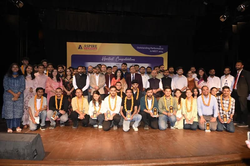 Large group photo on an auditorium stage at an Aspire felicitation ceremony: students wearing marigold garlands with faculty and guests in front of a congratulations backdrop