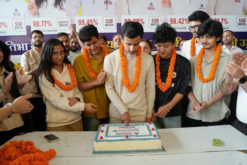 Garlanded Aspire students cutting a celebration cake surrounded by classmates, with result percentages displayed on a board behind them