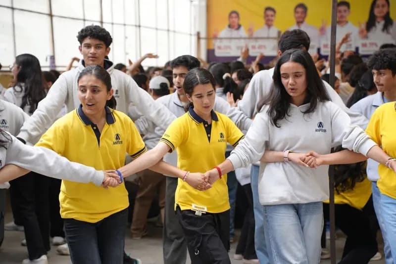 Aspire students in yellow T-shirts and grey hoodies holding hands and dancing in a circle at an institute celebration
